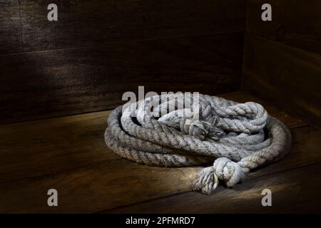 Ein altes, ausgefranstes, graues, aufgerolltes Seil auf einem rustikalen Holzambiente in sanfter, dunkler Stimmungsbeleuchtung mit Kopierbereich, das in einem Studio aufgenommen wurde Stockfoto