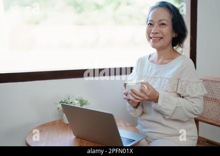 Porträt einer älteren Asiatin in moderner Pose, die Computerarbeit leistet und Kaffee trinkt. Stockfoto
