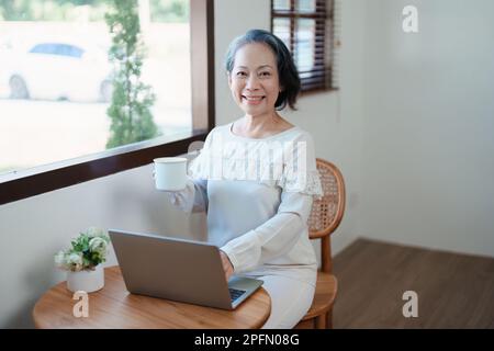 Porträt einer älteren Asiatin in moderner Pose, die Computerarbeit leistet und Kaffee trinkt. Stockfoto