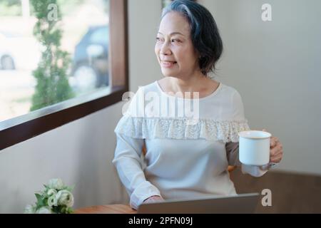 Porträt einer älteren Asiatin in moderner Pose, die Computerarbeit leistet und Kaffee trinkt. Stockfoto