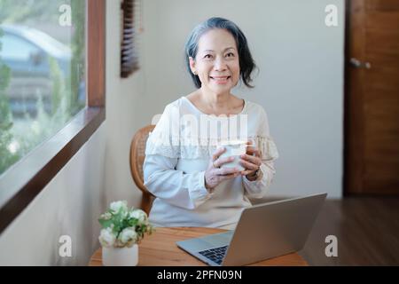Porträt einer älteren Asiatin in moderner Pose, die Computerarbeit leistet und Kaffee trinkt. Stockfoto