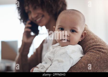 Mom alias Boss. Eine junge Frau, die ein Smartphone benutzt, während sie sich zu Hause um ihr niedliches Mädchen kümmert. Stockfoto