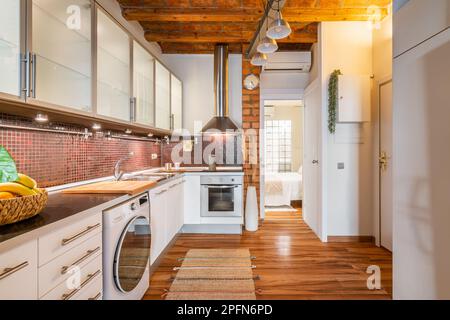 Stilvolle, gemütliche Küche mit modernen Geräten und Holzdecke mit Blick auf das offene Schlafzimmer mit Glasmauern. Konzept der lakonischen Konstruktion Stockfoto