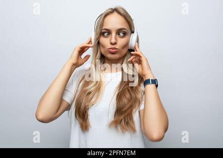 Eine junge Frau macht vor der Kamera ein Gesicht in weißen Kopfhörern auf weißem Hintergrund Stockfoto