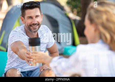 Glückliches junges Paar auf dem Campingplatz, das von Thermoskannen trinkt Stockfoto