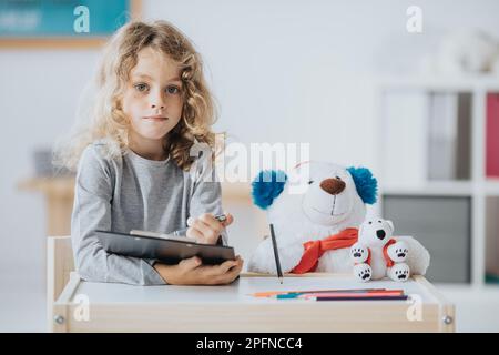 Ein junger Autist-Junge, der mit einem Teddybär an einem Schultisch sitzt Stockfoto