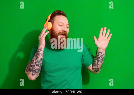Foto eines hübschen, fröhlichen Mannes mit Ingwerbart und stylischen T-Shirt-Kopfhörern, die zu Hause ein Lied singen, isoliert auf grünem Hintergrund Stockfoto