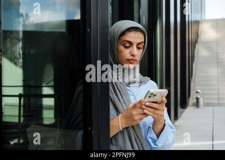 muslimische Frau mittleren Alters, die Hidschab trägt und im Freien mit dem Handy steht Stockfoto