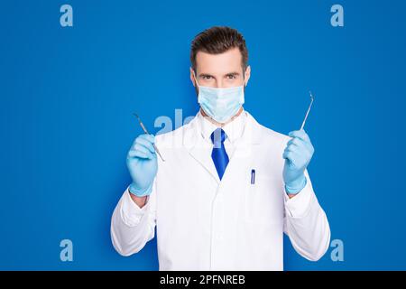 Porträt eines erfolgreichen gutaussehenden Zahnarztes in weißem Laborkittel mit blauer Krawatte, die Werkzeuge für die Zahnbehandlung mit den Händen hat, anschauen Stockfoto