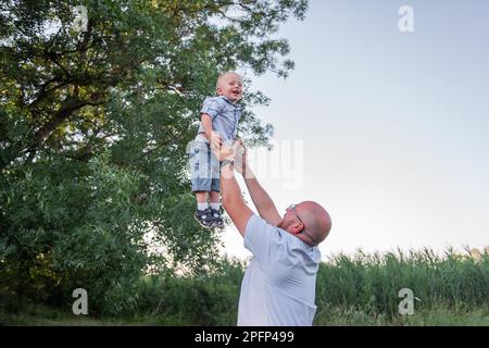 Ein Glatzkopf mit Brille wirft das Kind in die Luft. Vater in Jeans spielt, umarmt sich mit Sohn in der Natur außerhalb der Stadt. Der kleine Junge lacht, amüsiert sich Stockfoto