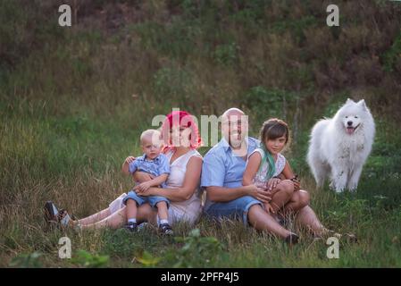 Porträt einer hellen, ungewöhnlichen, vielfältigen Familie in der Natur mit dem Samoyerten Hund. Reisen mit Haustieren. Kreative Leute schauen in die Kamera. Grüner botanischer Bac Stockfoto