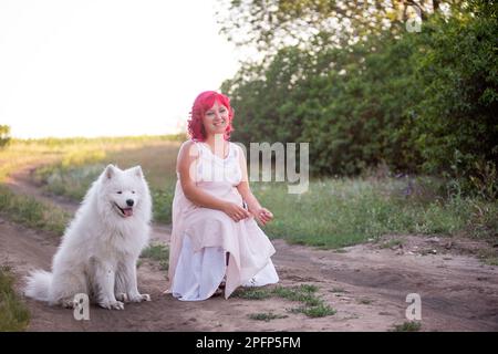 Leuchtende Diversity-Frau mit rosa Haaren wandert mit dem Samoyerten Hund im Wald entlang des Weges in den Sonnenuntergang. Kinderfreies Mädchen, Freundschaft mit Haustieren. Tr Stockfoto