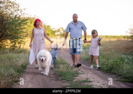 Samoierter Hund, der mit einer bunten Familie im Frühling auf den Wegen spaziert. Reisen mit Haustieren. Verschwommene Eltern halten Kinder an der Hand, Stockfoto