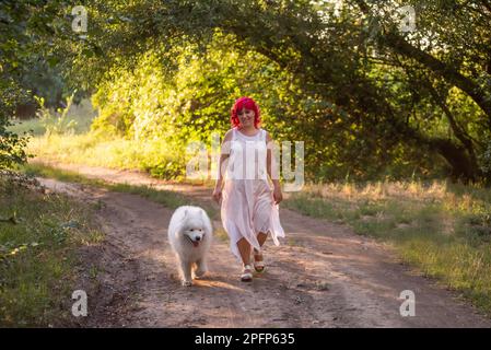 Leuchtende Diversity-Frau mit rosa Haaren wandert mit dem Samoyerten Hund im Wald entlang des Weges in den Sonnenuntergang. Kinderfreies Mädchen, Freundschaft mit Haustieren. Tr Stockfoto