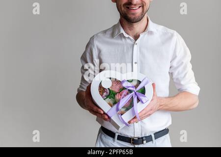 Ein gesichtsloser Mann hält die Form eines Herzens aus süßen Marshmallow-Blumen in den Händen. Geschenk mit lila Band. Zarte, zuckerfreie Rosen auf hellem Hintergrund. Sm Stockfoto