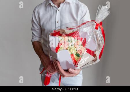 Ein gesichtsloser Mann hält einen Marshmallow-Blumenstrauß in den Händen. Geschenk in rotem Umschlag mit zarten, zuckerfreien Rosen auf hellem Hintergrund. Kleine Unternehmen Stockfoto