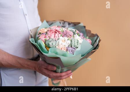 Ein gesichtsloser Mann hält einen Strauß süßer Marshmallow-Blumen in den Händen. Geschenk in pastellgrünem Umschlag mit zarten, zuckerfreien Rosen auf orangefarbenem Hintergrund. Sma Stockfoto