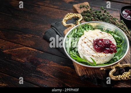 Gebackener Camembert Brie-Käse mit Preiselbeersauce und garniert mit Rucola-Salat in einer Pfanne. Holzhintergrund. Draufsicht. Speicherplatz kopieren. Stockfoto