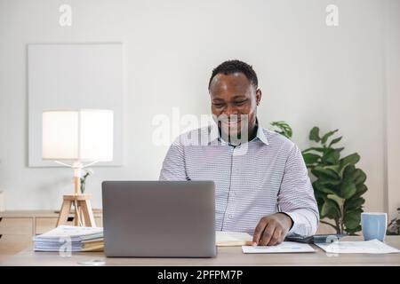 Fokussierter afrikanischer Geschäftsmann mit Dokumenten, der sich ein Notebook ansieht und recherchiert. Business Financial Analyst Konzept Stockfoto