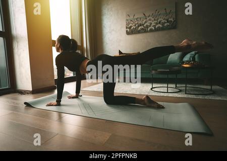 Frau übt zu Hause Yoga oder Workout in spezieller Sportkleidung auf Yogamatte. Ruhe- und Beschwichtigungskonzept. Eine Frau, die das Bein hochhält. Stockfoto
