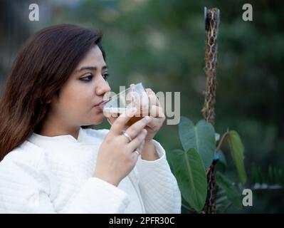 Junge Frauen, die eine transparente Teetasse halten und mit beiden Händen schwarzen Tee in einem Garten trinken, tragen eine weiße Wolljacke Stockfoto