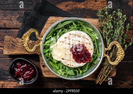 Gebackener Camembert Brie-Käse mit Preiselbeersauce und garniert mit Rucola-Salat in einer Pfanne. Holzhintergrund. Draufsicht. Stockfoto