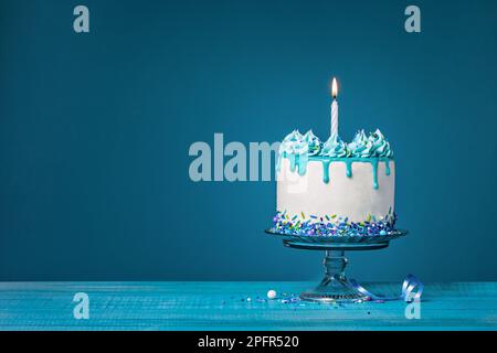 Weißer Tropfkuchen mit blauer Ganache, Streuseln und einer beleuchteten Geburtstagskerze auf blauem Hintergrund. Einfach und trendig. Speicherplatz kopieren. Stockfoto