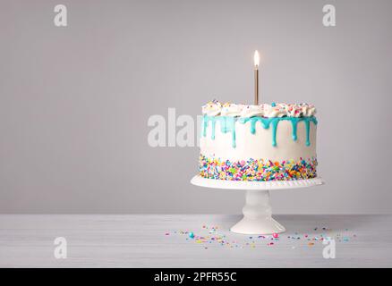 Vanille-Buttercreme-Geburtstagskuchen mit blauem Ganache-Tropf, brennender goldener Kerze und bunten Streuseln auf hellgrauem weißem Hintergrund. Speicherplatz kopieren. Stockfoto