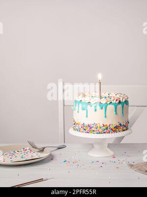 Geburtstagsparty mit Vanille-Buttercreme-Kuchen, türkisblauer Ganache-Tropf, brennender goldener Kerze und bunten Streuseln auf einem hellgrauen weißen Ba Stockfoto