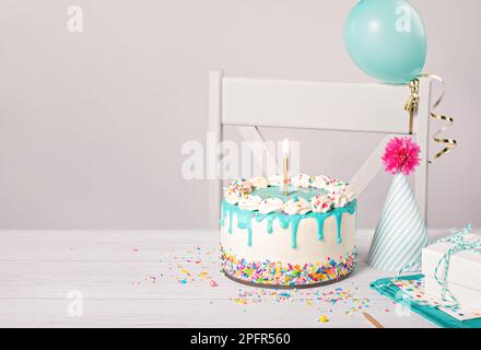 Geburtstagsparty mit einem Vanille-Buttercreme-Kuchen, blauer Ganache-Tropf, einer beleuchteten goldenen Kerze und bunten Streuseln auf einem hellgrauen weißen Ba Stockfoto