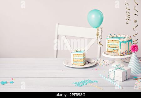 Happy Birthday Party Tisch Setting mit einem Vanille Buttercreme Konfetti Kuchen in Scheiben, blauer Ganache Tropf, auf hellgrauem weißem Hintergrund mit Ball Stockfoto