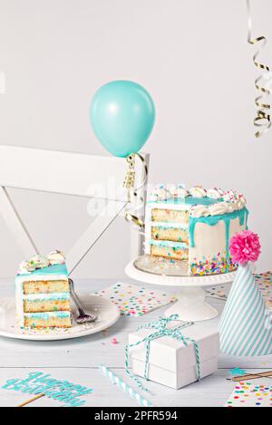 Happy Birthday Party Tisch Setting mit einem Vanille Buttercreme Konfetti Kuchen in Scheiben, blauer Ganache Tropf, auf hellgrauem weißem Hintergrund mit Ball Stockfoto