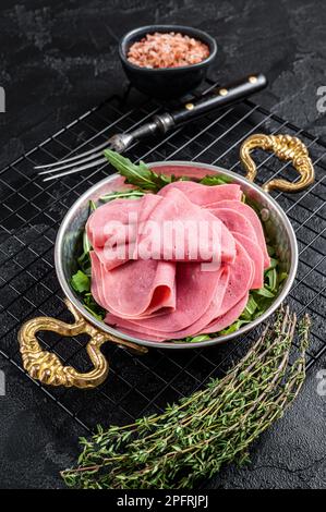 Dünne Schinkenscheiben in einer Pfanne mit Rucola-Salat. Schwarzer Hintergrund. Draufsicht. Stockfoto