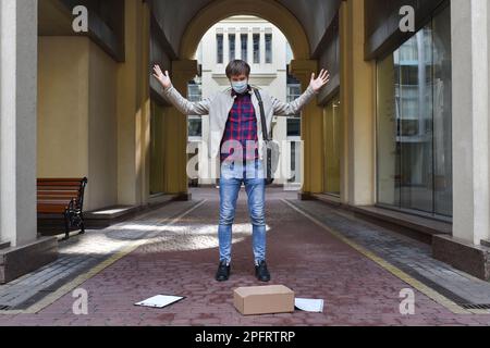 Emotionaler Mann in einer Schutzmaske vor dem Hintergrund eines modernen Gebäudes, von seinem Job gefeuert. Arbeitslosenproblem-Konzept. Eine Person, die leidet Stockfoto