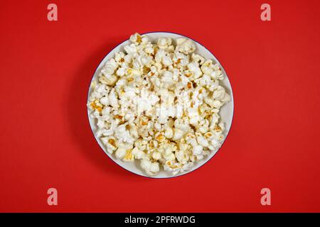 Popcorn auf einem Teller auf rotem Papierhintergrund. Nahaufnahme, Draufsicht Stockfoto