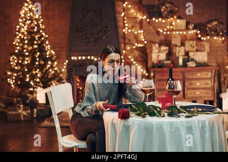 Junge enttäuschte Brünette sitzt am Tisch im Restaurant und hält die rote Schachtel mit einem Geschenk Stockfoto