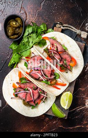 Carne Asada Tacos mit gegrilltem Steak, grüner Sauce, Jalapenos und Zwiebeln, mexikanische Küche. Dunkler Hintergrund. Draufsicht. Stockfoto