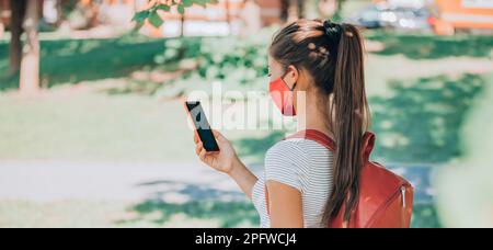 Student trägt Maske und Rucksack mit Handy-App für virtuelle Kurse an der Universität oder Universität. Schulfrau mit Handy in der Hand Stockfoto