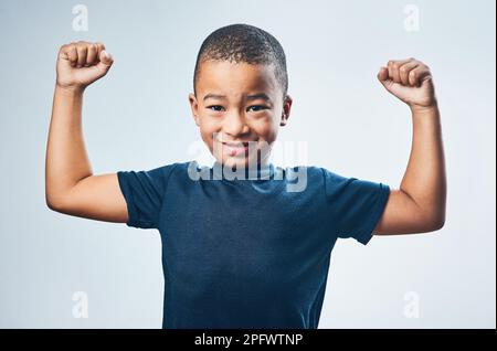 Ich bin jetzt ein großer Junge. Studioaufnahme eines süßen kleinen Jungen, der seine Muskeln vor grauem Hintergrund verbiegt. Stockfoto