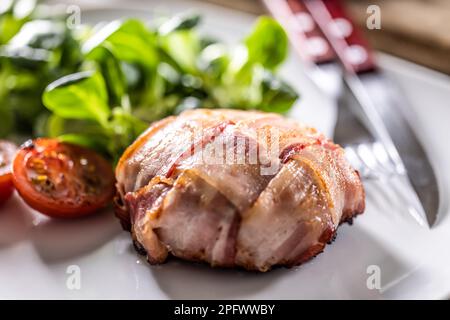 Fleisch in Speckscheiben eingewickelt, gebacken bis goldbraun und serviert mit Kirschtomaten und grünem Salat. Stockfoto