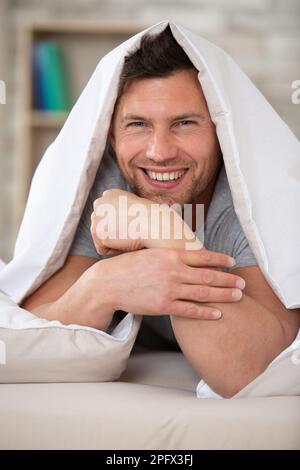 Glücklicher Mann, der sich zu Hause im Bett unter der Decke versteckt Stockfoto