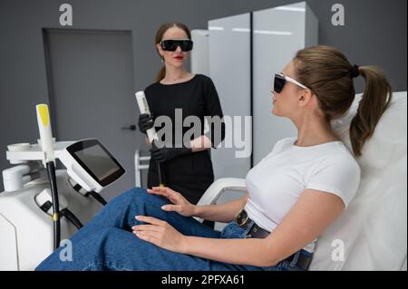 Eine Frau auf einer Couch in einer Brille, die sich einem Fotorejuvenationsverfahren unterzogen hat. Kosmetologe mit einem Patienten. Stockfoto