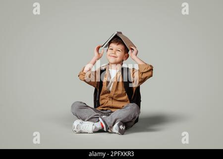 Der glückliche kleine europäische Junge mit Rucksack legt ein Buch auf den Kopf, sitzt auf dem Boden und lernt gern Stockfoto