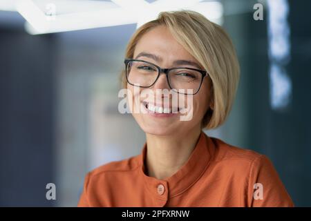 Nahaufnahme einer schönen blonden Geschäftsfrau im Büro, einer Brillenarbeiterin, die lächelt und in die Kamera schaut, Porträt einer glücklichen Frau. Stockfoto