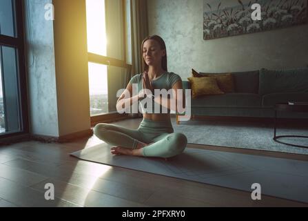 Frau übt Yoga und meditiert zu Hause mit geschlossenen Augen. Ruhe- und Beschwichtigungskonzept. Eine Frau, die in Lotus-Pose sitzt. Stockfoto