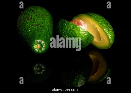 Ein ganzer und zwei Hälften, durchgeschnitten, reife grüne Avocados, auf schwarzem Hintergrund. Stockfoto