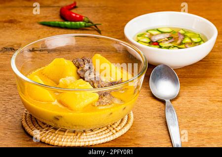 Traditionelle hausgemachte Kartoffeln, Chili, Rinderzwiebeln und Kokosmilch-Curry in transparenter Glasschale auf Bambusmatte mit Gurkenscheiben, Zwiebeln, V Stockfoto