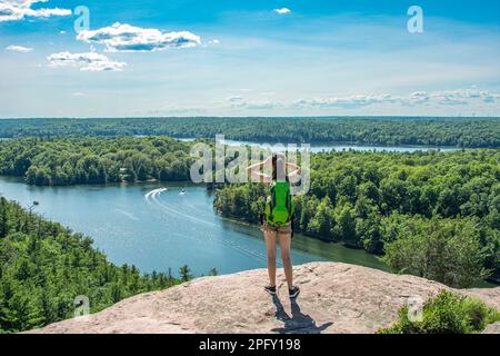 Ein Mädchen mit Rucksack steht hoch auf einem Felsen und bewundert den Blick auf die Seen und Wälder von oben Stockfoto