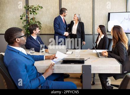 Eine Gruppe vielseitiger Geschäftsleute in formeller Kleidung, die am Tisch sitzen und Kollegen beim Händeschütteln während eines Geschäftstreffens in einem modernen Büro zuschauen Stockfoto