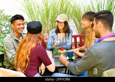 Eine Gruppe von fröhlichen, multiethnischen Freunden, die sich anschauen, während sie gemeinsam am Tisch frühstücken und Kaffee auf der Terrasse der Cafeteria im Freien trinken Stockfoto